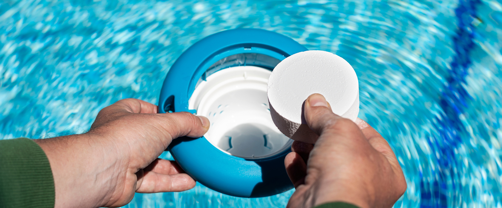 Hands placing a chlorine tablet into a blue floating dispenser in a swimming pool.