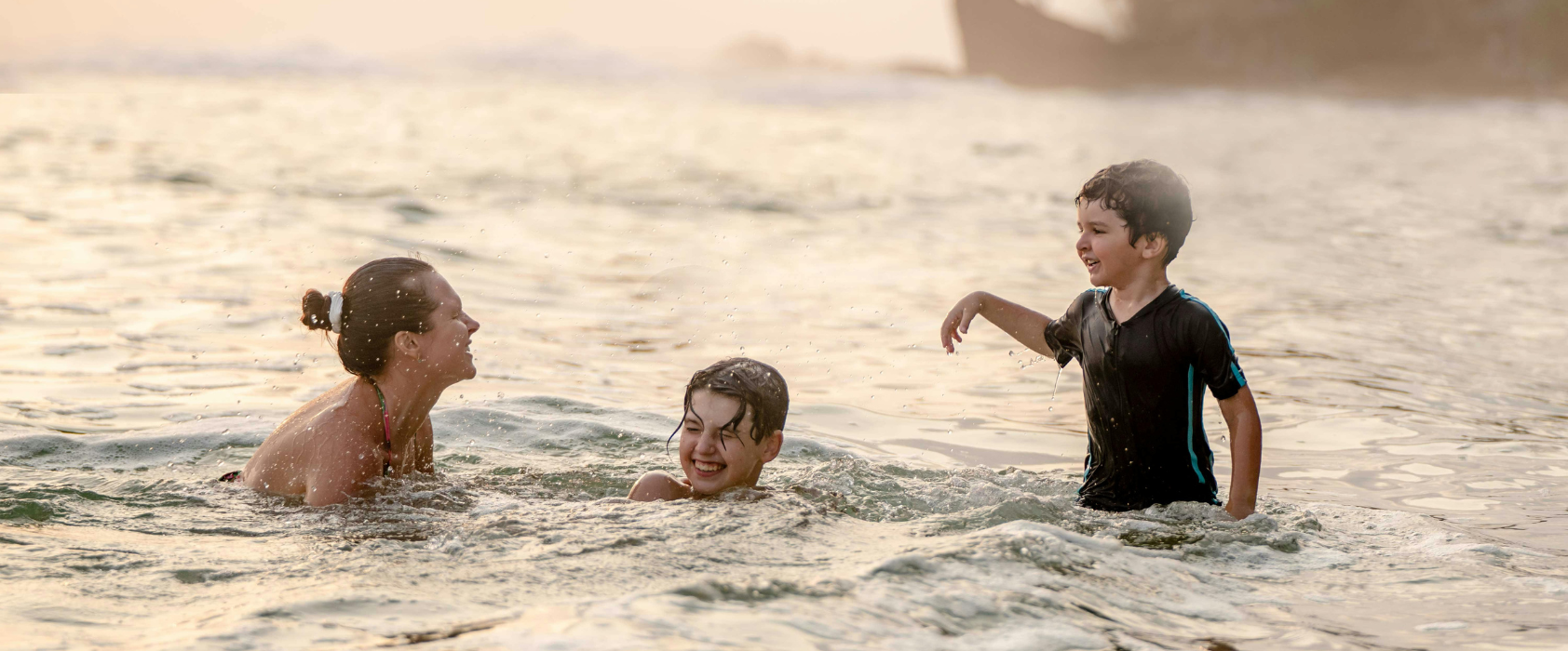 A woman and two children smiling and splashing together in shallow ocean water at sunset